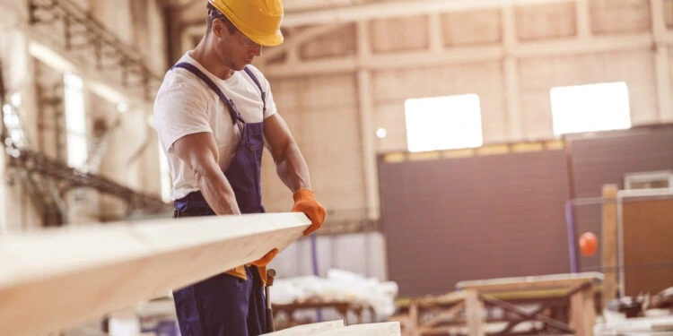 male-builder-carrying-wooden-plank-at-construction-2021-12-16-22-27-24-utc-scaled-750x375-1